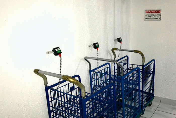 Iluminação Prédios e Condomínios imagem mostra três carrinhos de compras azuis empilhados e encostados em uma parede branca. Eles estão presos à parede por correntes de metal conectadas a dispositivos pretos e verdes (provavelmente um sistema de segurança ou travamento). No canto superior direito, há uma placa vermelha e branca