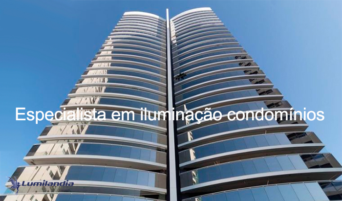 Iluminação Prédios e Condomínios A imagem mostra a entrada de um prédio residencial alto. Há um muro baixo com portão de madeira vertical e uma portaria com grandes janelas de vidro. O paisagismo é tropical, com palmeiras e canteiros de flores coloridas ao redor de um caminho pavimentado. O dia é ensolarado, com céu azul claros