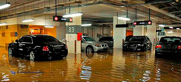 Iluminação Prédios e Condomínios A imagem mostra uma garagem de estacionamento subterrânea completamente alagada. A água suja, em tom marrom, cobre o chão e reflete as luzes do teto. Há vários carros de luxo estacionados, com a água atingindo a altura das rodas ou um pouco acima. Sinais de "NO ENTRY" (Não entre) estão visíveis